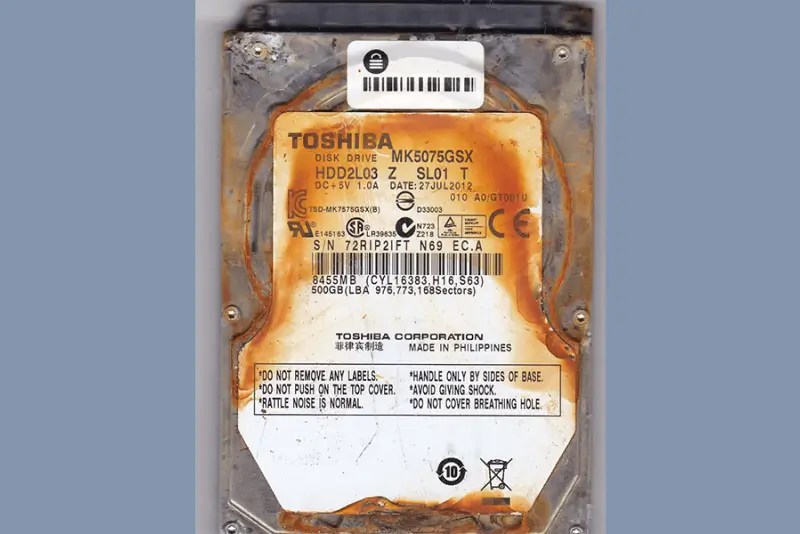 A rusted Toshiba 500GB hard drive with a label showing model MK5075GSX and date 27JUL2012. | TOSHIBA FU DISK DRIVE MK5075GSX HDD2L03 Z SL01 T DC+5V 1.0A TSD-MK7575GSX(B) SP E145163 LR39636 DATE: 27JUL2012 e. D33003 C N723 010 A0/GT001 AECE S/N 72RIP21FT N69 EC.A. 8455MB (CYL16383,H16, S63) 500GB (LBA 976,773,168Sectors) TOSHIBA CORPORATION 菲律宾制造 DO NOT REMOVE ANY LABELS *DO NOT PUSH ON THE TOP COVER. RATTLE NOISE IS NORMAL. MADE IN PHILIPPINES *HANDLE ONLY BY SIDES OF BASE AVOID GIVING SHOCK. *DO NOT COVER BREATHING HOLE. 10 ☑